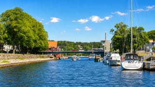 Sailboats moored along the Moss canal by a road bridge, with waterfront townhouses under a bright summer sky.