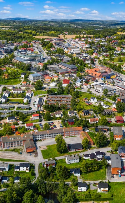 Aerial view of Mysen, Norway, a low-rise town center ringed by green farmland, forest and rolling hills under a blue sky.