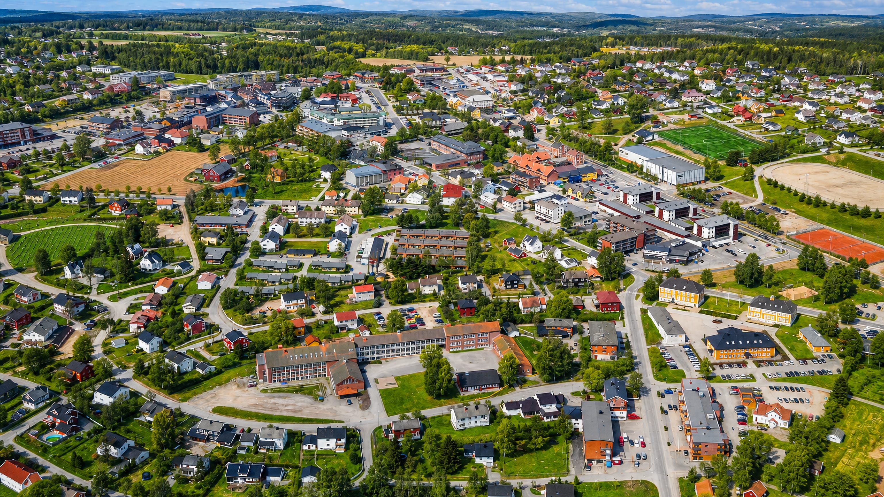 Aerial view of Mysen, Norway, a low-rise town center ringed by green farmland, forest and rolling hills under a blue sky.