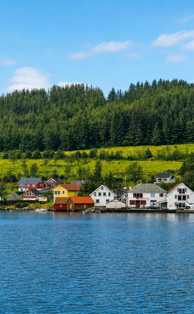 Colourful wooden houses line the calm fjord shore below forested green hills in the island village of Bokn, Norway.