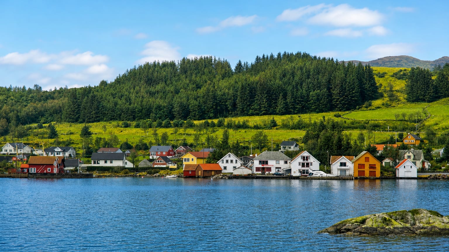Colourful wooden houses line the calm fjord shore below forested green hills in the island village of Bokn, Norway.