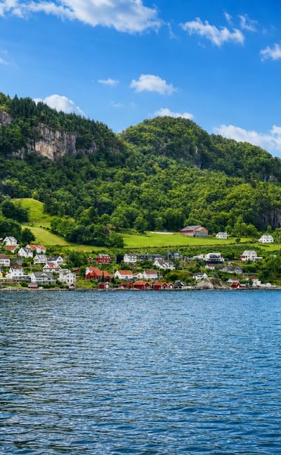 Hjelmelandsvågen's white and red waterfront houses below steep forested mountains on a fjord in Rogaland, Norway.