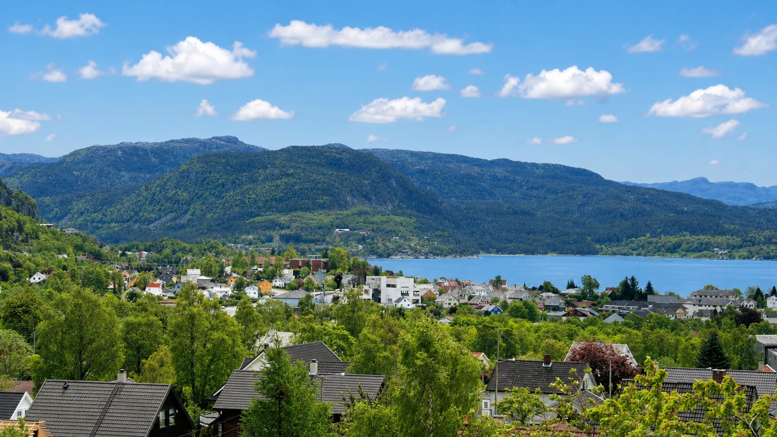 Jørpeland's houses spread along a fjord shore beneath forested Ryfylke mountains under a blue summer sky