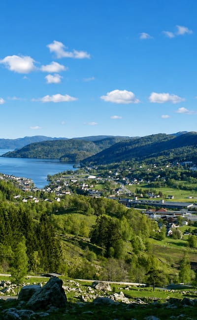 Elevated view over Moi, Norway, a village beside the long Lundevatnet lake in a forested valley ringed by green hills.