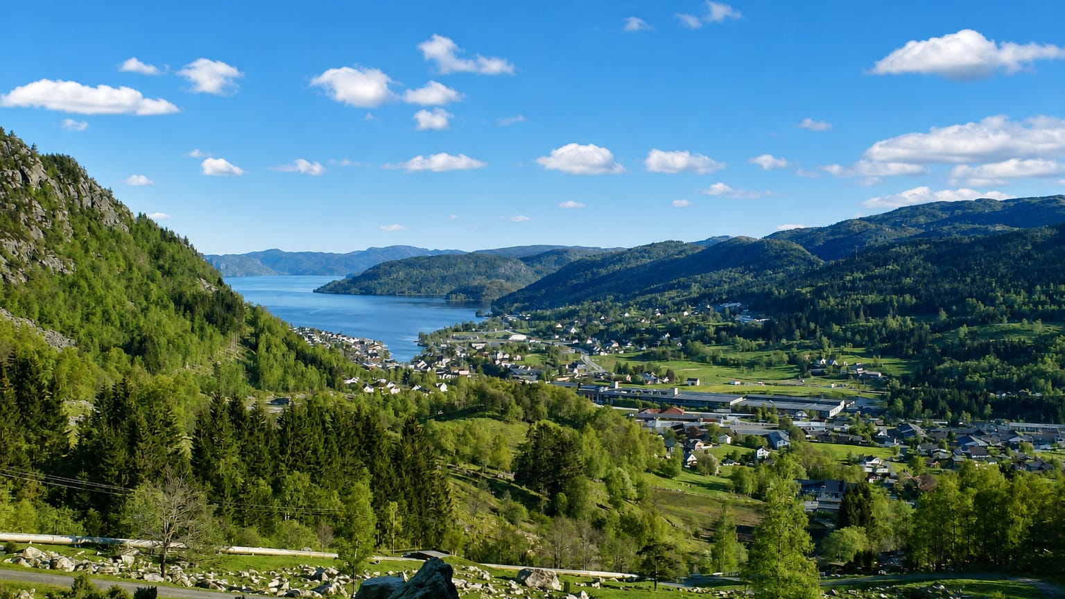 Elevated view over Moi, Norway, a village beside the long Lundevatnet lake in a forested valley ringed by green hills.