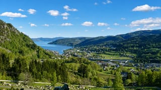 Elevated view over Moi, Norway, a village beside the long Lundevatnet lake in a forested valley ringed by green hills.