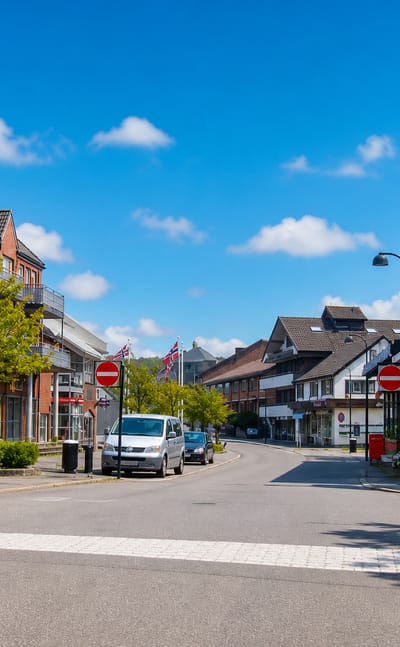 Quiet daytime shopping street in Sola, Norway, with red-brick apartments, parked cars and a pedestrian crossing under blue sky.