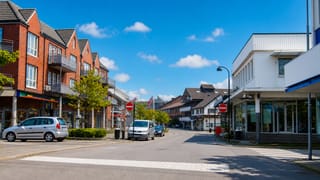 Quiet daytime shopping street in Sola, Norway, with red-brick apartments, parked cars and a pedestrian crossing under blue sky.
