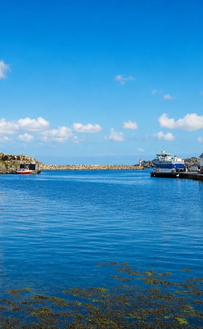 The island harbour of Utsira in Rogaland, Norway, with white timber houses, a ferry and rocky shoreline under a blue sky.
