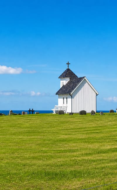 Varhaug's small white timber chapel and seaside graveyard on a grassy headland above the ocean under a clear sky.