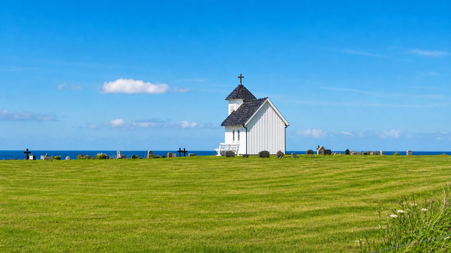 Varhaug's small white timber chapel and seaside graveyard on a grassy headland above the ocean under a clear sky.