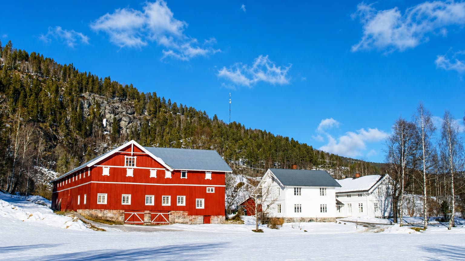 Red barn and white farmhouse on a snow-covered field below a forested hillside in Sauland, Norway.
