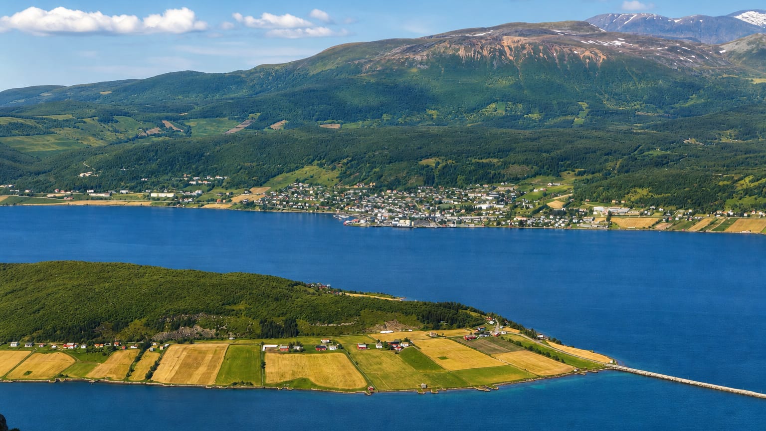 Elevated view of Borkenes village along a blue fjord with green farm fields and mountains, Troms, Norway.