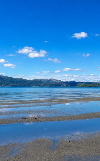Calm fjord at Brøstadbotn, Norway, with shallow tidal flats and low forested mountains under a bright blue summer sky.