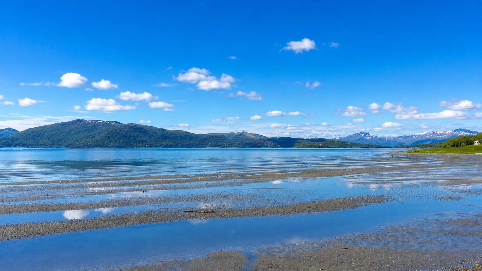 Calm fjord at Brøstadbotn, Norway, with shallow tidal flats and low forested mountains under a bright blue summer sky.