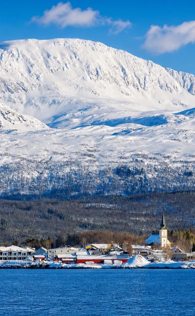 Snow-covered coastal village of Evenskjer, Norway, with a white church by a strait beneath a large snowy mountain.