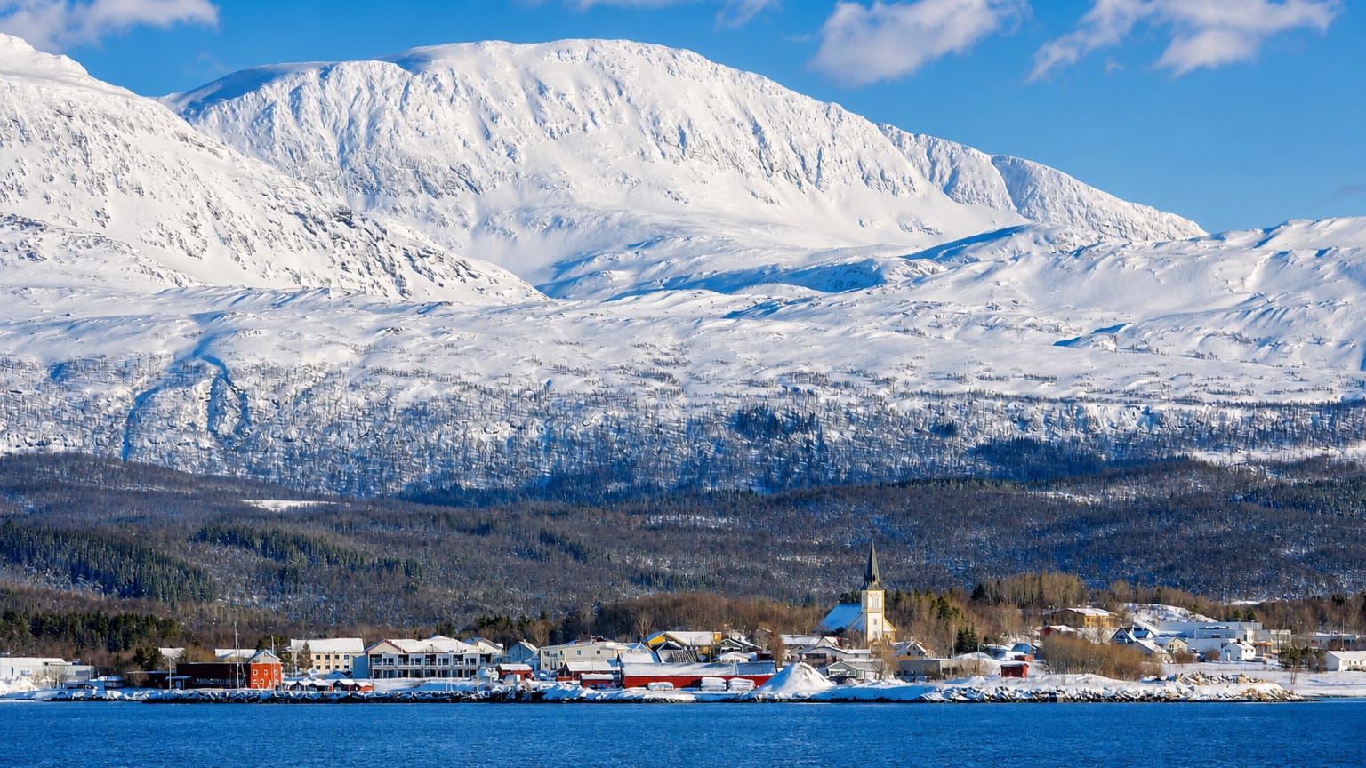 Snow-covered coastal village of Evenskjer, Norway, with a white church by a strait beneath a large snowy mountain.