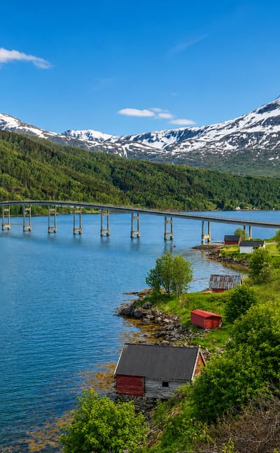 Long low bridge spanning the Gratangen fjord below snow-capped peaks, with red boathouses on the green shore