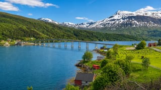 Long low bridge spanning the Gratangen fjord below snow-capped peaks, with red boathouses on the green shore