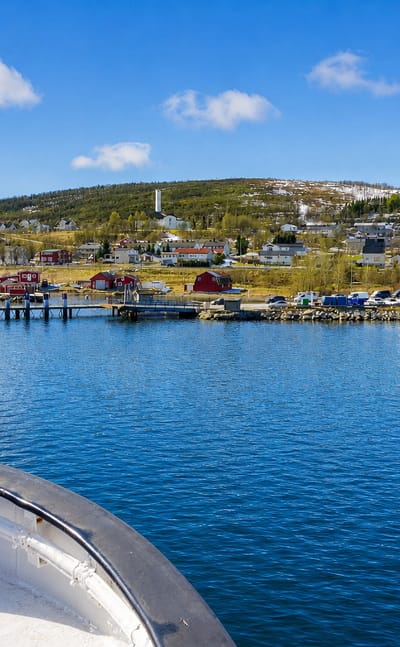 Hansnes coastal village and ferry quay across a calm blue fjord, with red houses and snow-dusted hills under a bright sky