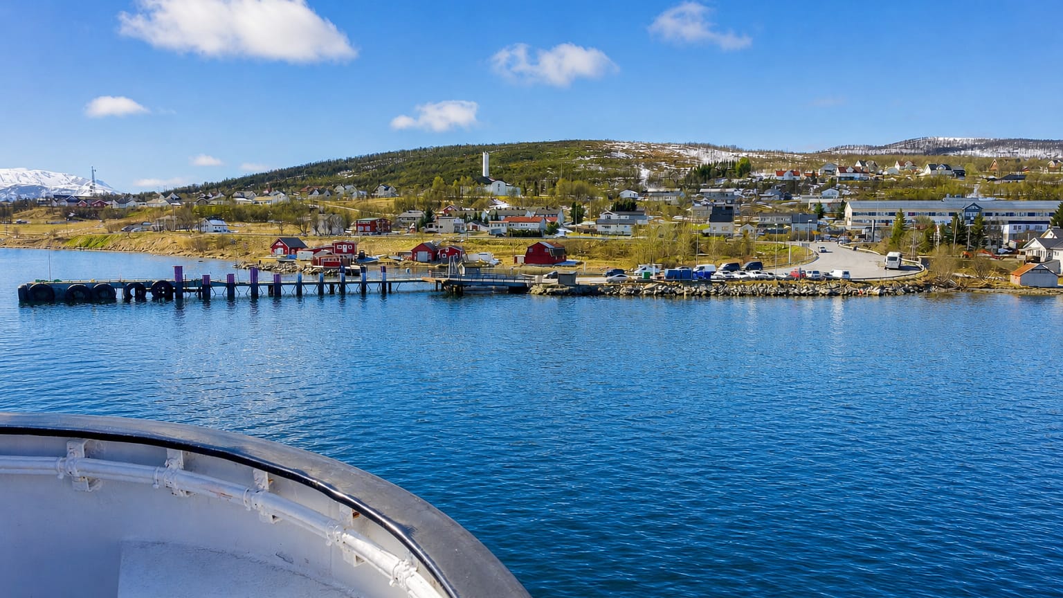 Hansnes coastal village and ferry quay across a calm blue fjord, with red houses and snow-dusted hills under a bright sky