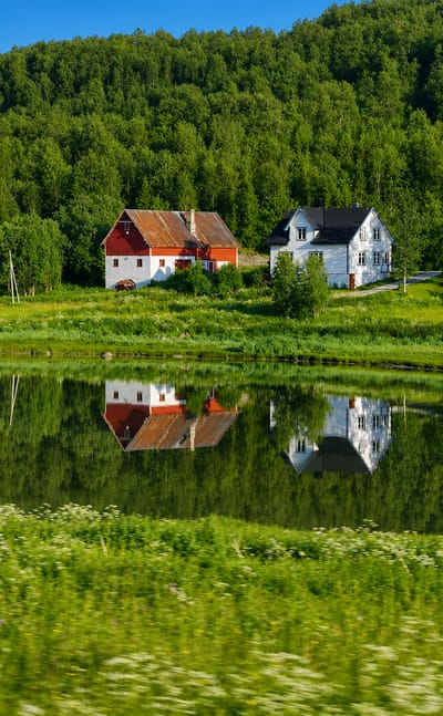 Red and white farmhouses reflected in still water amid green meadows at Sørreisa, Troms, Norway.