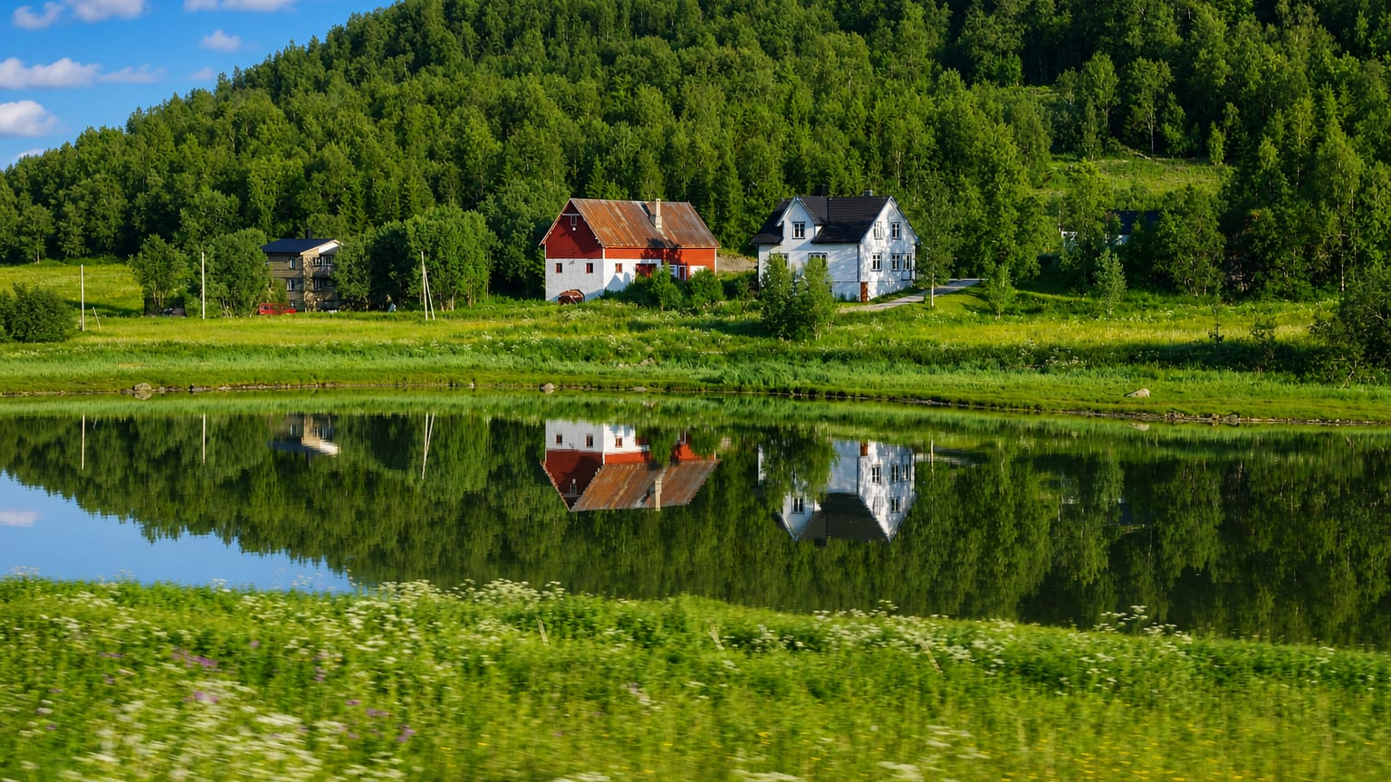 Red and white farmhouses reflected in still water amid green meadows at Sørreisa, Troms, Norway.