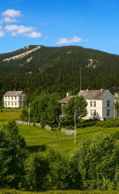 Whitewashed wooden farmhouses and a steepled white church among green pastures below forested hills in Berkåk, Norway.