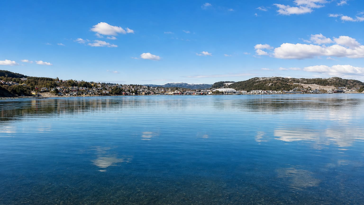 Botngård's low coastal settlement strung along the far shore of a calm fjord, with low hills behind under a blue sky.