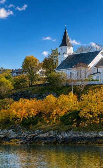 Fillan's white wooden hilltop church above the fjord, framed by autumn foliage and a rocky shoreline, Trøndelag, Norway