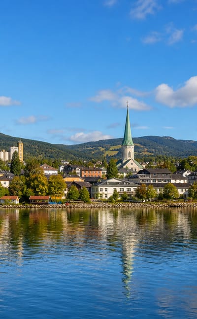 Grong waterfront with a green church spire, stone towers and forested hills mirrored in calm river water under a blue sky