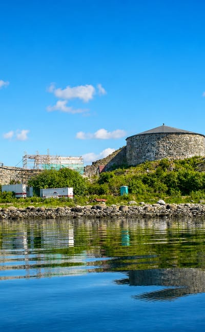 Stone fortress with a round tower on a green islet reflected in calm fjord water under a blue sky at Stjørdal, Norway
