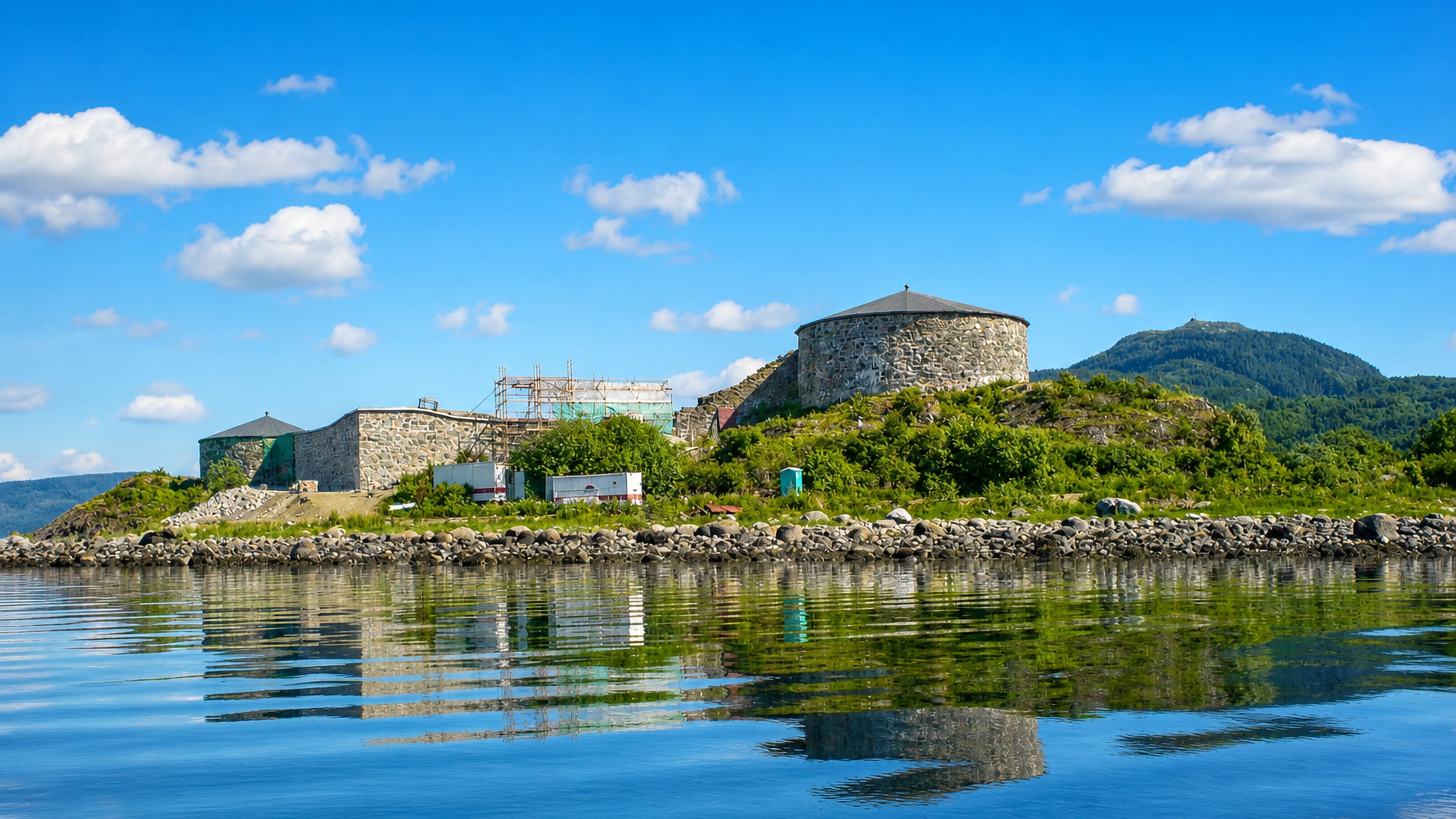 Stone fortress with a round tower on a green islet reflected in calm fjord water under a blue sky at Stjørdal, Norway