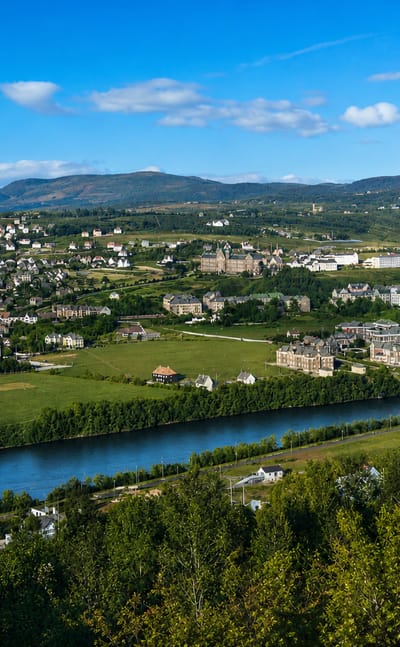 Elgeseter, Trondheim, Norway, seen across a river and green valley with hillside houses, a bridge and distant mountains.