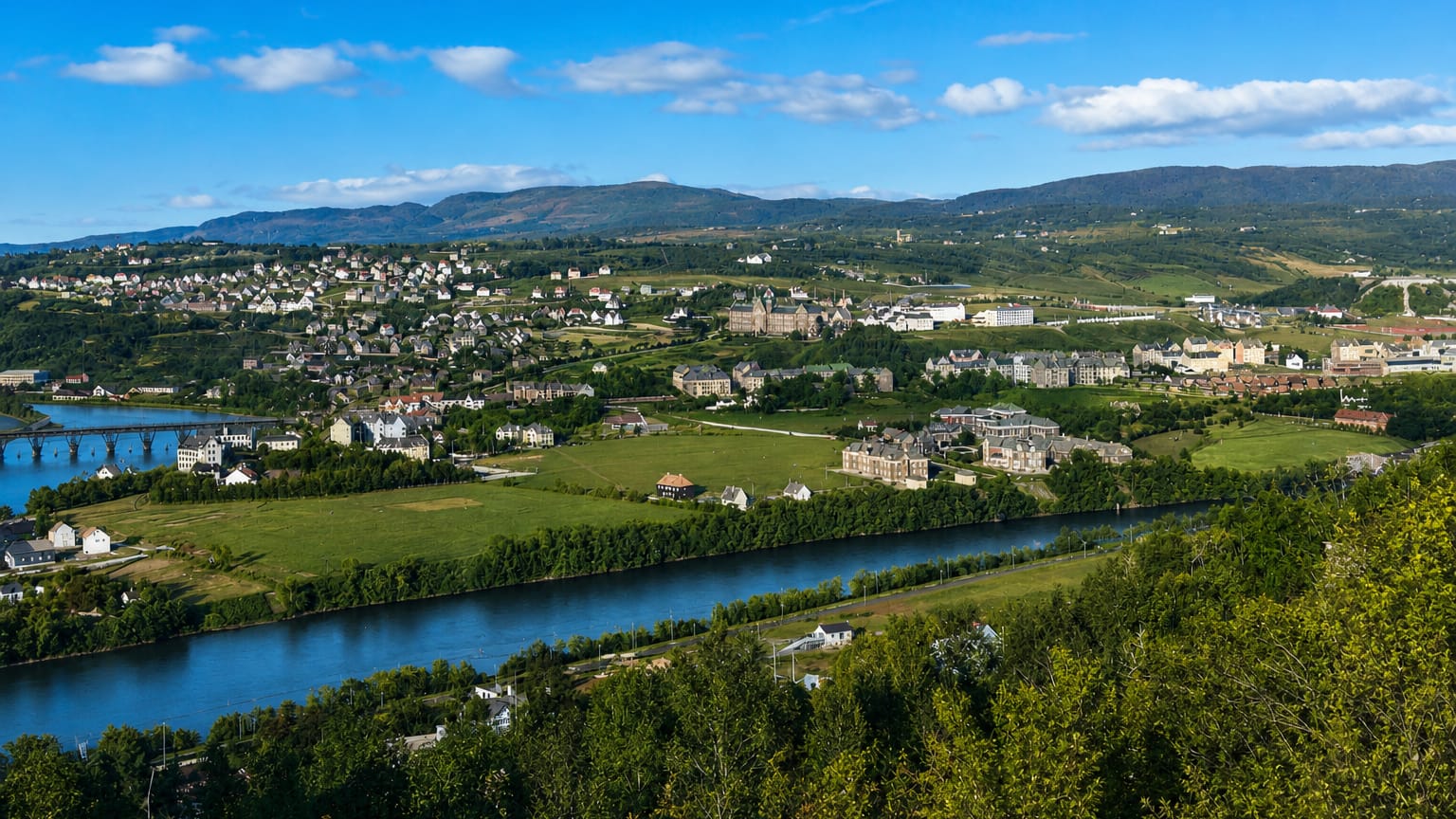 Elgeseter, Trondheim, Norway, seen across a river and green valley with hillside houses, a bridge and distant mountains.
