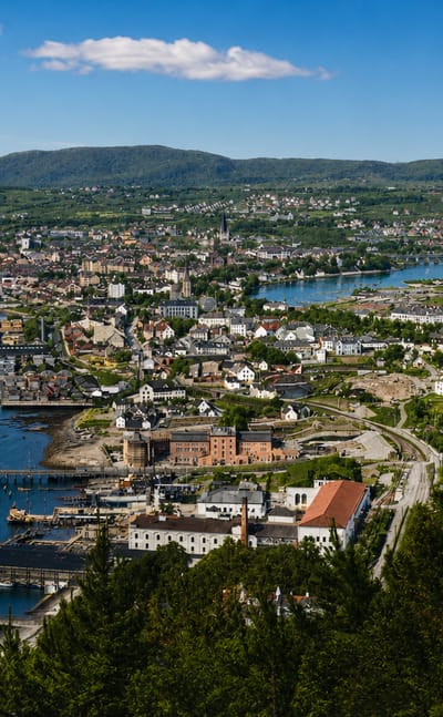 Aerial daytime view of Midtbyen, Trondheim — a coastal town with harbour piers, a winding river and forested hills.