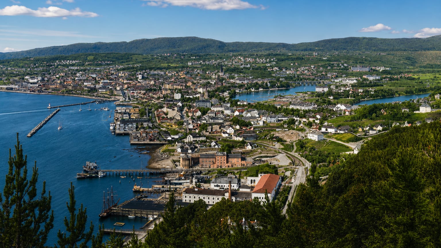 Aerial daytime view of Midtbyen, Trondheim — a coastal town with harbour piers, a winding river and forested hills.