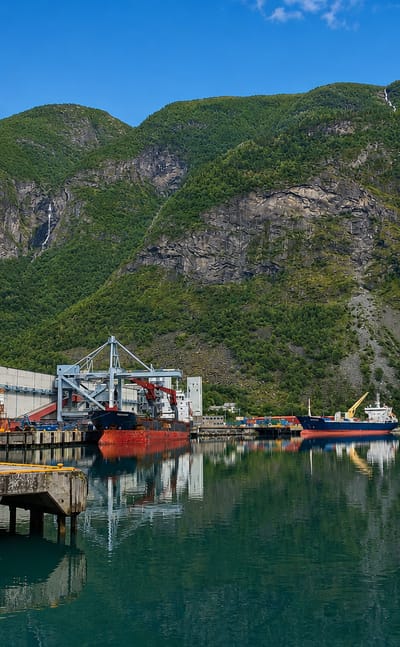 Årdalstangen's industrial fjord port with cargo ships at the dock, a waterfall and steep forested mountains at the head of Årdalsfjorden.
