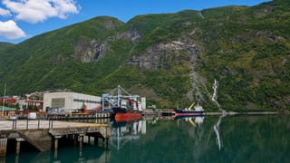 Årdalstangen's industrial fjord port with cargo ships at the dock, a waterfall and steep forested mountains at the head of Årdalsfjorden.