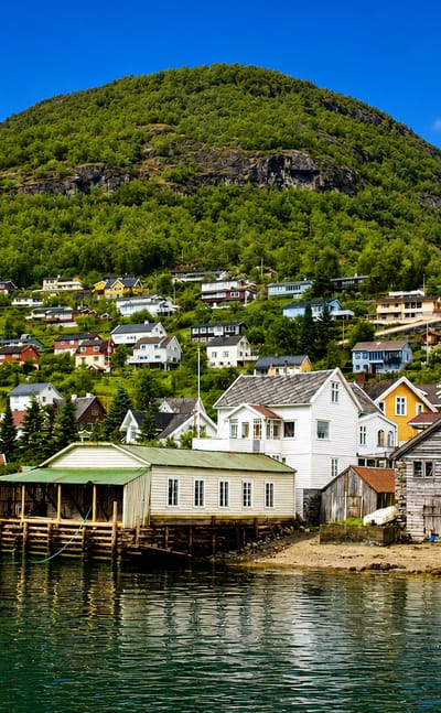 White wooden houses of Aurlandsvangen, Norway, on a green hillside above the calm fjord below a steep forested mountain