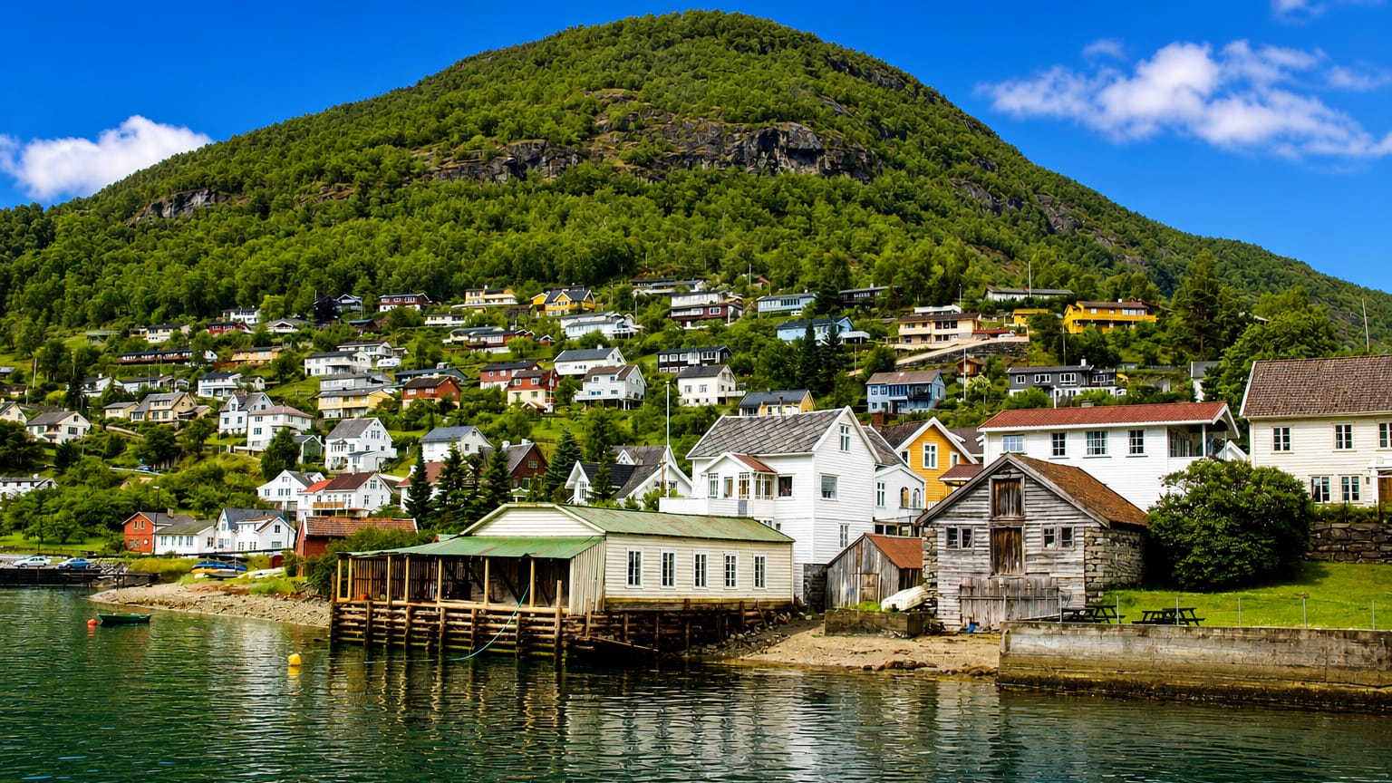 White wooden houses of Aurlandsvangen, Norway, on a green hillside above the calm fjord below a steep forested mountain