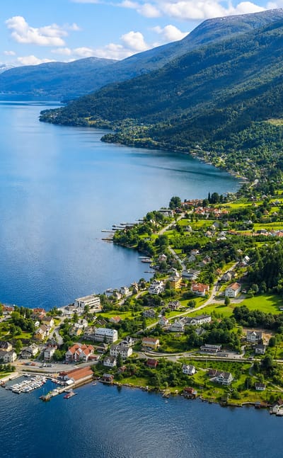 Aerial view of Balestrand village strung along the deep-blue Sognefjord, with a waterfront hotel and steep green mountain slopes