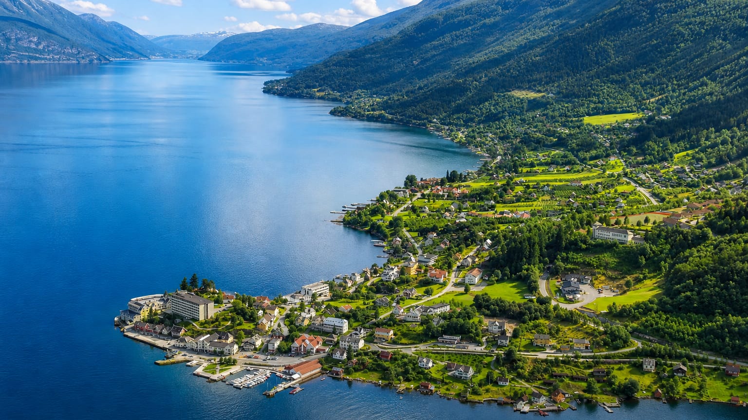 Aerial view of Balestrand village strung along the deep-blue Sognefjord, with a waterfront hotel and steep green mountain slopes