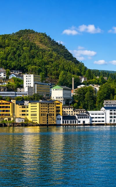 Fjordside factory buildings and hillside houses at Arna, Bergen, Norway, below green forested mountains under a blue sky