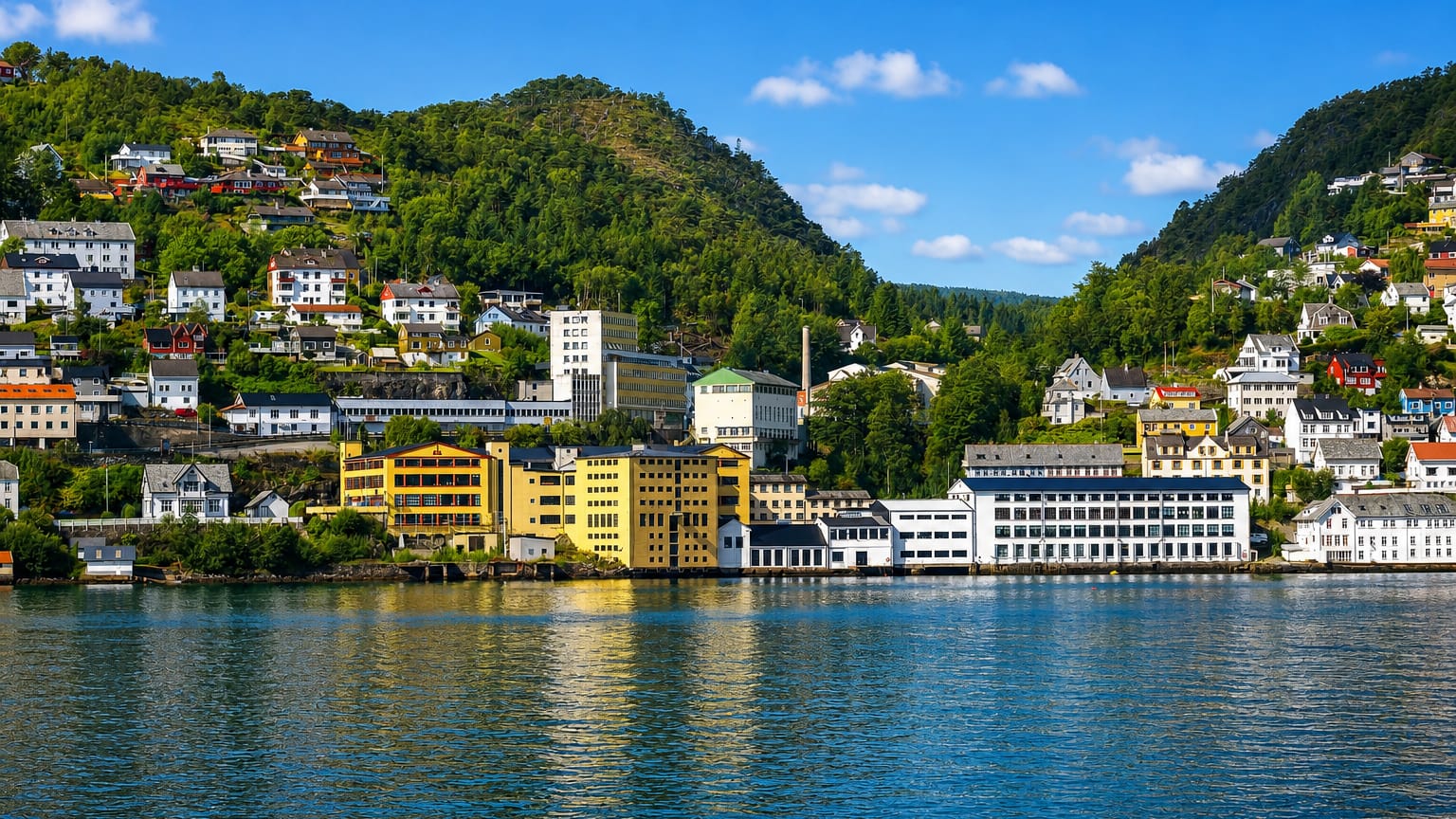 Fjordside factory buildings and hillside houses at Arna, Bergen, Norway, below green forested mountains under a blue sky
