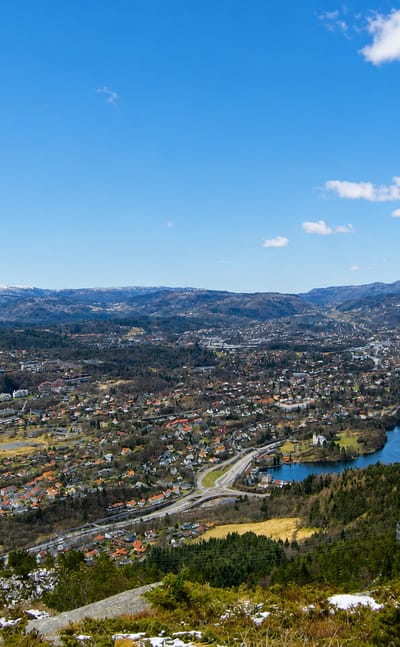 Elevated daytime panorama over Fana, Norway, a residential valley with a lake ringed by snow-capped hills south of Bergen.