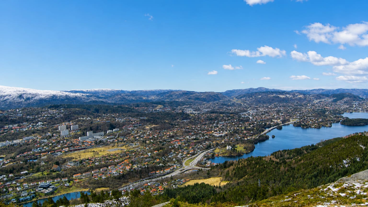 Elevated daytime panorama over Fana, Norway, a residential valley with a lake ringed by snow-capped hills south of Bergen.