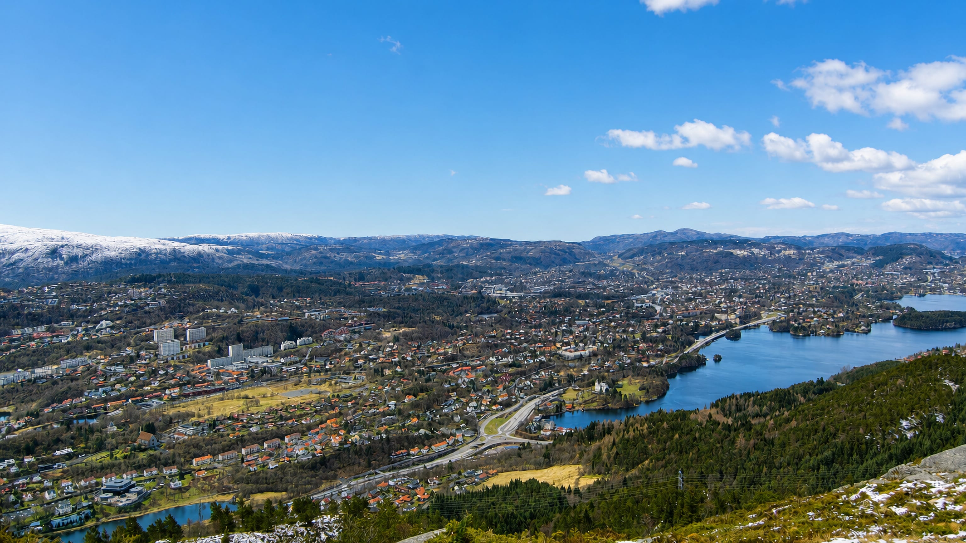 Elevated daytime panorama over Fana, Norway, a residential valley with a lake ringed by snow-capped hills south of Bergen.