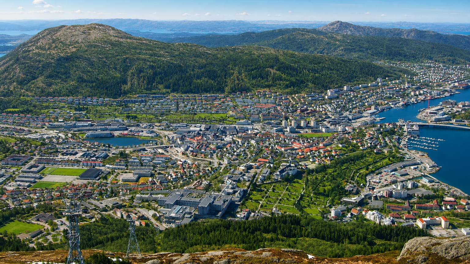 Elevated daytime view over Kronstad, Bergen, low-rise housing in a green valley framed by forested hills and fjord inlets.