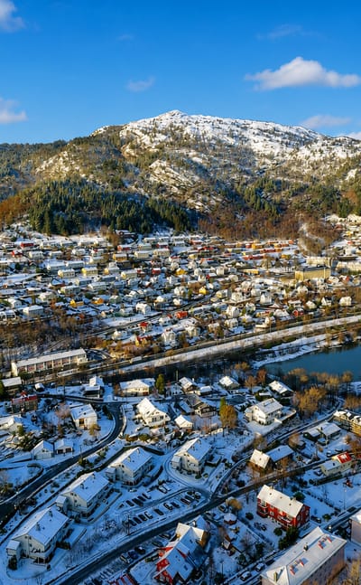 Snow-covered residential neighborhood of Minde, Norway, beside a small lake below a forested mountain under a blue sky.